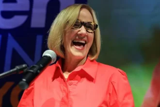 A smiling woman speaks at a microphone during an event, wearing glasses and a bright red shirt.