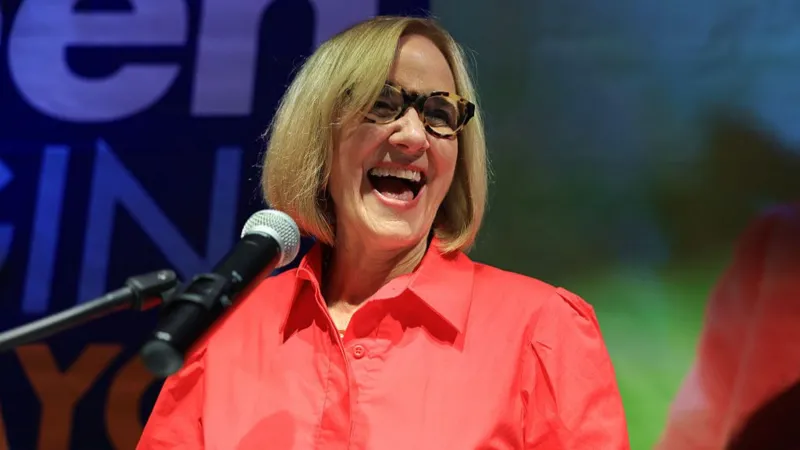 A smiling woman speaks at a microphone during an event, wearing glasses and a bright red shirt.
