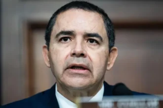 A middle-aged man wearing a dark suit speaks into a microphone at a formal hearing, with a wooden panel background behind him.