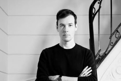 Black-and-white portrait of a fashion designer standing on a staircase with arms crossed, reflecting a moment of creative transition at Leonard Paris.