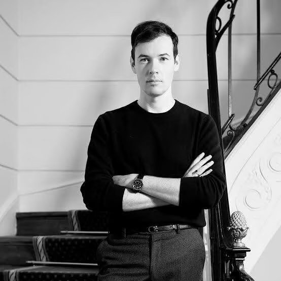 Black-and-white portrait of a fashion designer standing on a staircase with arms crossed, reflecting a moment of creative transition at Leonard Paris.