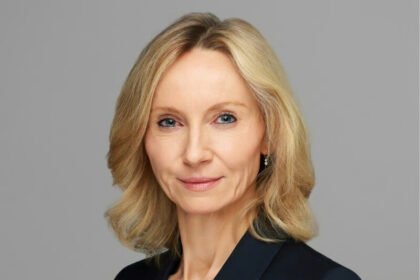 A professional portrait of a blonde businesswoman in a dark blazer, photographed against a grey background