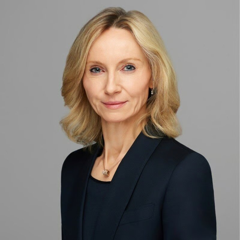 A professional portrait of a blonde businesswoman in a dark blazer, photographed against a grey background