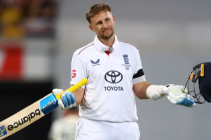 An England cricketer in a white Test uniform stands on the field holding his bat in one hand and his helmet in the other, giving a small shrug with a calm, almost playful expression after completing a milestone innings.