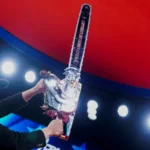 A man on stage raises a large chrome chainsaw under bright event lights.