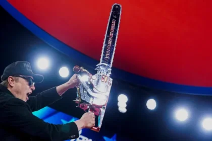 A man on stage raises a large chrome chainsaw under bright event lights.