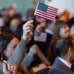 A crowd of people sit together at a ceremony, many of them holding small American flags, with one person in the foreground raising a flag in the air.