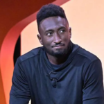 A man in a black long-sleeve shirt sits on stage against an orange backdrop, looking thoughtful during a tech-related discussion.