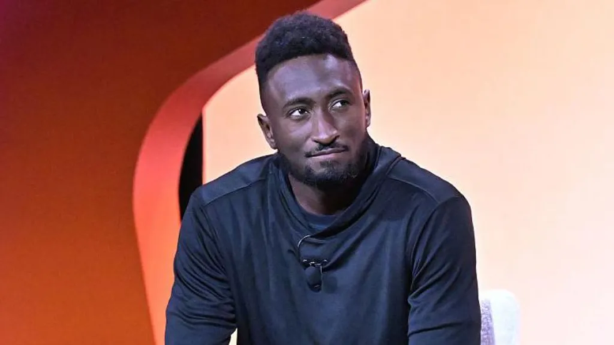 A man in a black long-sleeve shirt sits on stage against an orange backdrop, looking thoughtful during a tech-related discussion.