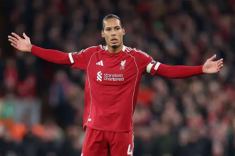 A soccer player wearing a red Liverpool FC kit, identified as Virgil van Dijk, stands on the pitch with his arms stretched out to his sides in a gesture of confusion or exasperation. He is wearing the captain's armband and his jersey has the standard chartered sponsor logo. The background is a slightly blurred, dark stadium crowd.