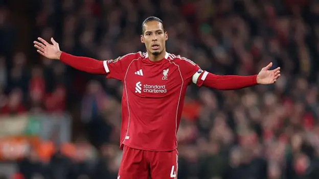A soccer player wearing a red Liverpool FC kit, identified as Virgil van Dijk, stands on the pitch with his arms stretched out to his sides in a gesture of confusion or exasperation. He is wearing the captain's armband and his jersey has the standard chartered sponsor logo. The background is a slightly blurred, dark stadium crowd.