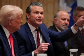A group of men in formal suits sit at a conference table. The man in the middle is speaking passionately and pointing forward, while the man to his left listens closely. Two other men sit on the right, watching the discussion.