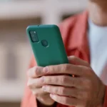 A close-up view of a person's hands holding a green smartphone with a rear fingerprint sensor. The person is wearing a burnt orange button-down shirt over a white t-shirt, and the background features blurred shelves.