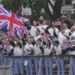 A large group of athletes, wearing matching light blue and white uniforms, are waving small Union Jack flags and cheering on a boat or platform, with a large Union Jack banner behind them.