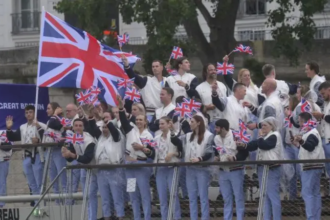 A large group of athletes, wearing matching light blue and white uniforms, are waving small Union Jack flags and cheering on a boat or platform, with a large Union Jack banner behind them.