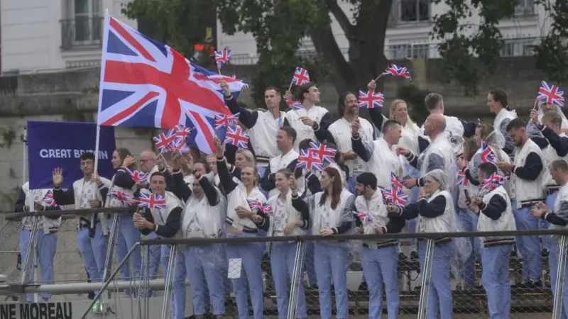 A large group of athletes, wearing matching light blue and white uniforms, are waving small Union Jack flags and cheering on a boat or platform, with a large Union Jack banner behind them.