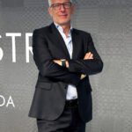 A Confindustria Moda representative stands confidently in a dark suit with arms crossed in front of a patterned grey backdrop.