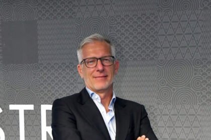 A Confindustria Moda representative stands confidently in a dark suit with arms crossed in front of a patterned grey backdrop.