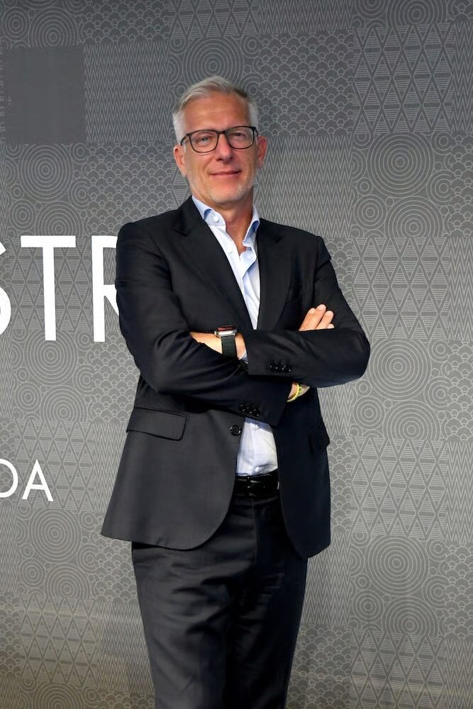 A Confindustria Moda representative stands confidently in a dark suit with arms crossed in front of a patterned grey backdrop.
