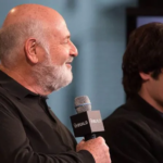 An elderly filmmaker holding a microphone stands beside a younger man during a public event, amid news surrounding the arrest of a son in a high-profile Hollywood homicide case.