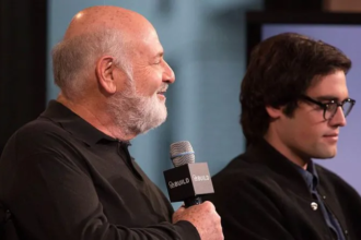 An elderly filmmaker holding a microphone stands beside a younger man during a public event, amid news surrounding the arrest of a son in a high-profile Hollywood homicide case.