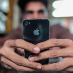 A person holds an iPhone with both hands, focusing on the screen while sitting indoors. The back of the phone with the Apple logo is clearly visible, and the background appears softly blurred.
