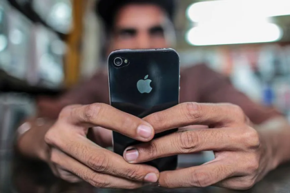 A person holds an iPhone with both hands, focusing on the screen while sitting indoors. The back of the phone with the Apple logo is clearly visible, and the background appears softly blurred.