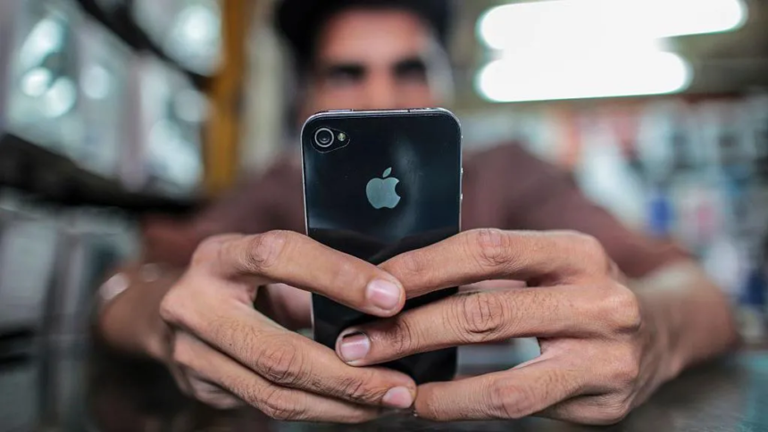 A person holds an iPhone with both hands, focusing on the screen while sitting indoors. The back of the phone with the Apple logo is clearly visible, and the background appears softly blurred.
