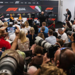 A wide shot of a press conference with three Formula 1 drivers seated on a stage against an Abu Dhabi Grand Prix backdrop, surrounded by many photographers and journalists holding cameras.