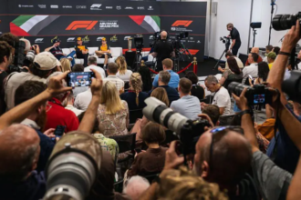 A wide shot of a press conference with three Formula 1 drivers seated on a stage against an Abu Dhabi Grand Prix backdrop, surrounded by many photographers and journalists holding cameras.
