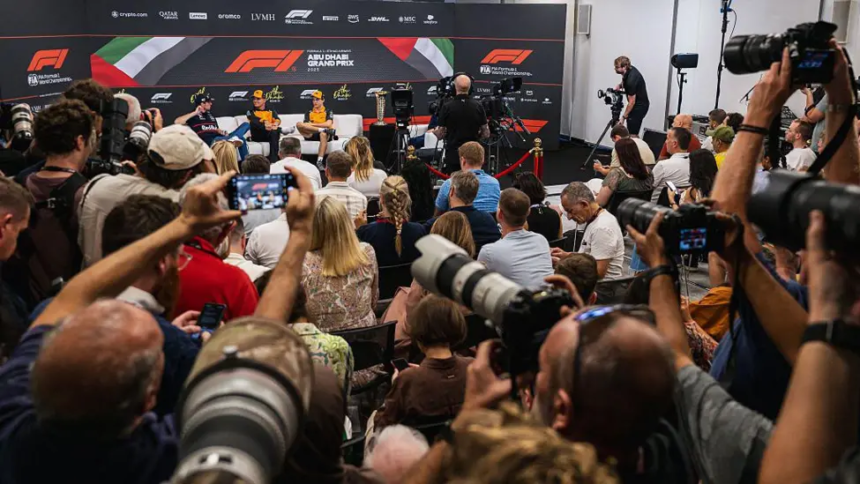 A wide shot of a press conference with three Formula 1 drivers seated on a stage against an Abu Dhabi Grand Prix backdrop, surrounded by many photographers and journalists holding cameras.