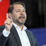 Senator Ruben Gallego speaks at a press conference about legislation to block President Trump from invading Greenland, with U.S. and Greenland flags in the background.
