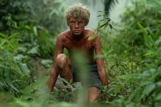 Lord of the Flies TV Review: A Dark, Gripping Take on a Classic Survival Story ★★★★☆ Jack Thorne returns to familiar ground with his new television adaptation of Lord of the Flies, but this time the scale feels bigger and the impact deeper. After the global success of Adolescence, which he co created with Stephen Graham, Thorne turns to a literary giant. He adapts Lord of the Flies by William Golding for a bold four part series that feels both faithful and refreshingly new. A Classic Story, Told With New Urgency The story remains the same at its core. A group of schoolboys survive a plane crash and find themselves stranded on a remote island. At first, they try to create order. Soon, fear and power struggles take over. Civilization slips away. Violence follows. While the original novel works as a broad allegory about society and human nature, this adaptation sharpens its emotional focus. Thorne balances two layers carefully. On one level, the series plays as a tense survival thriller. On another, it becomes a chilling study of group psychology and moral collapse. Unlike Adolescence, which centered on one troubled boy, this series explores collective behavior. It asks how ordinary children can drift toward brutality when authority disappears. Strong Direction and Striking Visual Style Director Marc Munden brings intensity to every frame. He uses unusual camera angles and close ups to create unease. Nature is not just background here. Insects crawl. Ants swarm. The island feels alive and indifferent. The colors stand out. Bright reds and harsh greens give scenes a dreamlike, almost nightmarish feel. The music, composed by Cristobal Tapia de Veer, adds to the tension. The score rumbles beneath key moments, heightening dread without overpowering the story. Each episode follows a different character’s point of view. This structure builds intimacy. It helps viewers understand how fear spreads and how loyalties shift. Deeper Character Focus Without Losing the Core Message Thorne adds small glimpses into the boys’ lives before the crash. These flashbacks provide context, especially for Jack and others. Still, the real strength of the story lies in its archetypes. Ralph stands out as the elected leader. He tries to act responsibly. Yet he also mocks Piggy to gain approval. That flaw makes him human. It also shows how easily decency bends under pressure. Although the series centers on children, it is not meant only for young viewers. It raises hard questions about power, conformity, and morality. Families who watch together will likely leave with strong opinions and difficult conversations. Final Verdict This adaptation of Lord of the Flies succeeds because it respects the novel’s core themes while giving them fresh energy. It feels unsettling, thoughtful, and visually bold. Jack Thorne proves once again that stories about youth can expose uncomfortable truths about society as a whole. Rating: ★★★★☆
