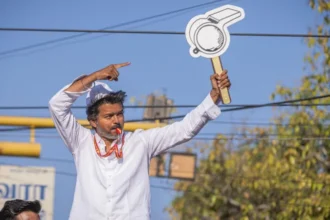 Political uncertainty in Tamil Nadu after actor Vijay’s party falls short of majority Historic win but no clear mandate C Joseph Vijay, a well known film star turned politician, has led his newly formed party Tamilaga Vettri Kazhagam (TVK) to a major breakthrough in Tamil Nadu’s state election. The party won 108 seats in the 234 member assembly, becoming the single largest party. However, it still fell 10 seats short of the 118 needed for a majority. This result created a hung assembly, leaving the state without a clear path to form a stable government. Coalition talks begin amid uncertainty After the results, the Indian National Congress, holding five seats, announced its support for TVK. Even so, Vijay still needs at least five more legislators to reach the majority mark. He met Tamil Nadu Governor Rajendra Vishwanath Arlekar and formally staked claim to form the government. The governor later stated that TVK has not yet shown proof of majority support in the assembly. Reports suggest the governor has asked Vijay to demonstrate backing from at least 118 members before any invitation to form government is considered. Debate over constitutional process TVK leaders and allies have criticized the governor’s approach, arguing that the single largest party should normally be given a chance to prove majority on the assembly floor. Legal and constitutional experts remain divided. Some say the governor is acting to ensure political stability, while others believe established convention supports inviting the largest party first and allowing a floor test later. A shift in Tamil Nadu politics TVK’s rise has been compared to earlier political disruptions in the state, especially when MG Ramachandran broke away to form AIADMK and later became chief minister. For decades, Tamil Nadu politics has been dominated by DMK and AIADMK. This election broke that pattern, with TVK emerging as the strongest single party and defeating the ruling DMK. Possible outcomes still open In the past few days, political discussions have intensified. There is even speculation about unlikely alliances between long time rivals. However, analysts say Vijay may still secure support from smaller parties and independents to reach the required number. The final outcome now depends on post election negotiations, not just the vote results.