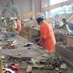 Robots Step In as Waste Industry Struggles to Hire Staff Recycling Plant Faces Labour Shortage Challenges A busy recycling facility in Rainham, East London, is dealing with ongoing staff shortages in a tough working environment. The site is operated by Sharp Group, a family run waste management company. The plant processes around 280,000 tonnes of mixed recycling every year. Workers sort materials on fast moving conveyor belts where items like shoes, broken electronics, and construction waste pass by constantly. The job is physically demanding and noisy. Dust is present throughout the facility, making conditions even harder for staff. Safety risks are also a concern in this sector, which reports higher injury and illness rates compared to many other industries. High Turnover Continues to Pressure Operations Keeping staff has become a major challenge. The facility reports an annual turnover rate of around 40 percent. Line supervisor Ken Dordoy explains that many workers struggle with the pace. He notes that pickers are constantly working as the belt never stops for long, which leads to frequent staff replacements. To manage fatigue, workers rotate between different sorting tasks every short interval, and the belt is occasionally paused to give brief breaks. Robots Enter Recycling Workflows To address staffing issues, the company is testing automation. A humanoid robot called Alpha is being trained to assist with sorting waste. The robot has been developed by RealMan Robotics in China and is being adapted for recycling operations by TeknTrash Robotics in the United Kingdom. Unlike traditional industrial machines, this robot is designed to move in a human like way. Developers believe this approach allows it to work within existing facilities without major redesigns. Training the Robot to Sort Waste Alpha is still in early development and is undergoing training. It is learning how to identify different items on the conveyor belt and how to pick them up correctly. A worker uses a virtual reality headset to demonstrate sorting techniques. These movements are recorded to help train the robot. According to TeknTrash founder and chief executive Al Costa, the training process focuses on two main skills. The first is recognising materials on the belt. The second is learning how to handle and lift them safely and efficiently.