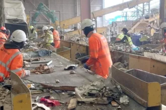 Robots Step In as Waste Industry Struggles to Hire Staff Recycling Plant Faces Labour Shortage Challenges A busy recycling facility in Rainham, East London, is dealing with ongoing staff shortages in a tough working environment. The site is operated by Sharp Group, a family run waste management company. The plant processes around 280,000 tonnes of mixed recycling every year. Workers sort materials on fast moving conveyor belts where items like shoes, broken electronics, and construction waste pass by constantly. The job is physically demanding and noisy. Dust is present throughout the facility, making conditions even harder for staff. Safety risks are also a concern in this sector, which reports higher injury and illness rates compared to many other industries. High Turnover Continues to Pressure Operations Keeping staff has become a major challenge. The facility reports an annual turnover rate of around 40 percent. Line supervisor Ken Dordoy explains that many workers struggle with the pace. He notes that pickers are constantly working as the belt never stops for long, which leads to frequent staff replacements. To manage fatigue, workers rotate between different sorting tasks every short interval, and the belt is occasionally paused to give brief breaks. Robots Enter Recycling Workflows To address staffing issues, the company is testing automation. A humanoid robot called Alpha is being trained to assist with sorting waste. The robot has been developed by RealMan Robotics in China and is being adapted for recycling operations by TeknTrash Robotics in the United Kingdom. Unlike traditional industrial machines, this robot is designed to move in a human like way. Developers believe this approach allows it to work within existing facilities without major redesigns. Training the Robot to Sort Waste Alpha is still in early development and is undergoing training. It is learning how to identify different items on the conveyor belt and how to pick them up correctly. A worker uses a virtual reality headset to demonstrate sorting techniques. These movements are recorded to help train the robot. According to TeknTrash founder and chief executive Al Costa, the training process focuses on two main skills. The first is recognising materials on the belt. The second is learning how to handle and lift them safely and efficiently.