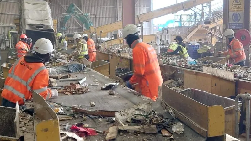 Robots Step In as Waste Industry Struggles to Hire Staff Recycling Plant Faces Labour Shortage Challenges A busy recycling facility in Rainham, East London, is dealing with ongoing staff shortages in a tough working environment. The site is operated by Sharp Group, a family run waste management company. The plant processes around 280,000 tonnes of mixed recycling every year. Workers sort materials on fast moving conveyor belts where items like shoes, broken electronics, and construction waste pass by constantly. The job is physically demanding and noisy. Dust is present throughout the facility, making conditions even harder for staff. Safety risks are also a concern in this sector, which reports higher injury and illness rates compared to many other industries. High Turnover Continues to Pressure Operations Keeping staff has become a major challenge. The facility reports an annual turnover rate of around 40 percent. Line supervisor Ken Dordoy explains that many workers struggle with the pace. He notes that pickers are constantly working as the belt never stops for long, which leads to frequent staff replacements. To manage fatigue, workers rotate between different sorting tasks every short interval, and the belt is occasionally paused to give brief breaks. Robots Enter Recycling Workflows To address staffing issues, the company is testing automation. A humanoid robot called Alpha is being trained to assist with sorting waste. The robot has been developed by RealMan Robotics in China and is being adapted for recycling operations by TeknTrash Robotics in the United Kingdom. Unlike traditional industrial machines, this robot is designed to move in a human like way. Developers believe this approach allows it to work within existing facilities without major redesigns. Training the Robot to Sort Waste Alpha is still in early development and is undergoing training. It is learning how to identify different items on the conveyor belt and how to pick them up correctly. A worker uses a virtual reality headset to demonstrate sorting techniques. These movements are recorded to help train the robot. According to TeknTrash founder and chief executive Al Costa, the training process focuses on two main skills. The first is recognising materials on the belt. The second is learning how to handle and lift them safely and efficiently.