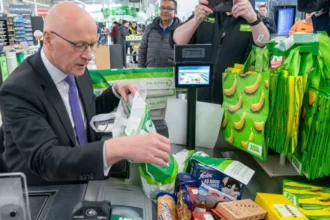 Swinney Defends Food Price Cap Plan Ahead of First Minister Vote SNP Leader Promises Relief for Families Facing Rising Food Costs John Swinney has defended his proposal to cap the prices of essential food items as he prepares for a key vote to remain Scotland’s first minister. He said the policy focuses on helping families manage the ongoing cost of living crisis rather than creating political conflict with the UK government. Speaking ahead of the parliamentary vote, Swinney explained that many households continue to struggle with the rising cost of groceries. According to him, the government has a responsibility to make healthy and basic food items more affordable for the public. Under the proposed SNP plan, major supermarkets in Scotland would have to limit the prices of up to 50 everyday products. These products could include milk, eggs, rice, cheese, and other household essentials. The policy aims to reduce pressure on low and middle income families who are finding it difficult to keep up with food expenses. Supermarkets and Retailers Raise Concerns The proposal has already sparked debate among retailers and business groups. The Scottish Retail Consortium criticized the plan and called it unrealistic. Retail representatives warned that smaller stores could struggle to compete because the price cap would mainly target large supermarket chains. Business leaders also argued that limiting prices could create challenges for suppliers and retailers. However, Swinney responded by saying the government would work closely with supermarkets, producers, and other stakeholders before finalizing the legislation. He stressed that public support remains his main concern because many people across Scotland continue to face financial hardship. Swinney added that discussions with retailers would play an important role in making the policy practical and effective. UK Law Could Create Challenges The SNP leader admitted that introducing the food pricing law may require cooperation with the UK government. Experts believe the proposal could conflict with the UK Internal Market Act 2020, which was introduced after Brexit to prevent trade barriers between Scotland, England, Wales, and Northern Ireland. Since the SNP no longer holds a parliamentary majority, Swinney acknowledged that he would need support from opposition parties in Holyrood as well as discussions with ministers in Westminster. Despite criticism from political opponents, Swinney rejected claims that the policy was designed to trigger a political dispute with London. Instead, he insisted that his goal is to support people who are struggling to afford basic shopping needs. Additional Promises in SNP Manifesto Alongside the food pricing proposal, the SNP has also promised to introduce a £2 cap on bus fares and expand childcare services across Scotland. The party says these measures would help reduce financial pressure on working families and improve access to affordable public services. Swinney believes the combined policies would provide direct support to households dealing with inflation and rising living costs.