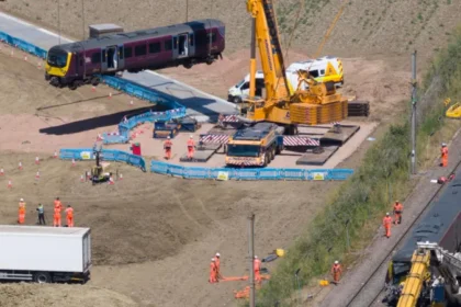 Train Manager Praised After Deadly East Midlands Railway Crash Near Bedford Emergency response at the crash scene A train manager has been praised for his quick thinking and bravery after a serious collision involving two East Midlands Railway services near Bedford. The crash happened on Friday at around 17:15 BST. One driver, Shaun Burton, died in the collision. Around 100 passengers were injured. Passengers said the train manager was hurt but still focused on safety. He used the radio to alert control rooms and helped coordinate an emergency response while checking on passengers. Heroic actions prevented further risk Representatives from the RMT Union said the train manager acted fast to prevent a second incident. He saw that one train had derailed and could block another line. He warned control staff immediately. He also placed circuit clips on the track system. This action forced signals to turn red and stopped other trains from entering the area. He then contacted the signaller to shut down movement on the line. Union officials said these actions may have stopped another train from hitting the damaged carriages. Witness accounts from passengers Passenger Mareks Grabovskis described the moment of impact. He said he had been in the last carriage after changing seats at the final moment. He said the crash threw him forward and he briefly lost consciousness. When he woke up, he saw injured passengers and panic inside the carriage. He also praised the train manager for staying calm and helping others even while injured himself. Tributes to driver Shaun Burton The driver who died, Shaun Burton, has been described as a respected member of staff. Local officials, including Blake Stephenson, shared condolences and highlighted the impact on his family and community. Sarah Conboy, leader of Huntingdonshire District Council, said Burton had served as a councillor between 2018 and 2022 and was well respected locally. Major rail disruption and repair work Recovery work is ongoing across the crash site. Officials say the damage is severe. A 600 metre section of track needs full replacement. Overhead electric lines also require removal and rebuilding. Heavy cranes are being used to lift damaged train carriages. Rail teams expect disruption between London and Bedford to continue for at least a week. Officials say access to the site is difficult, which is slowing recovery work. Medical response and passenger care A doctor who was travelling on one of the trains suffered fractures but still helped treat injured passengers at the scene. Emergency services later took the doctor to hospital. Passengers arriving at stations said staff have been providing support and updates as services slowly return.