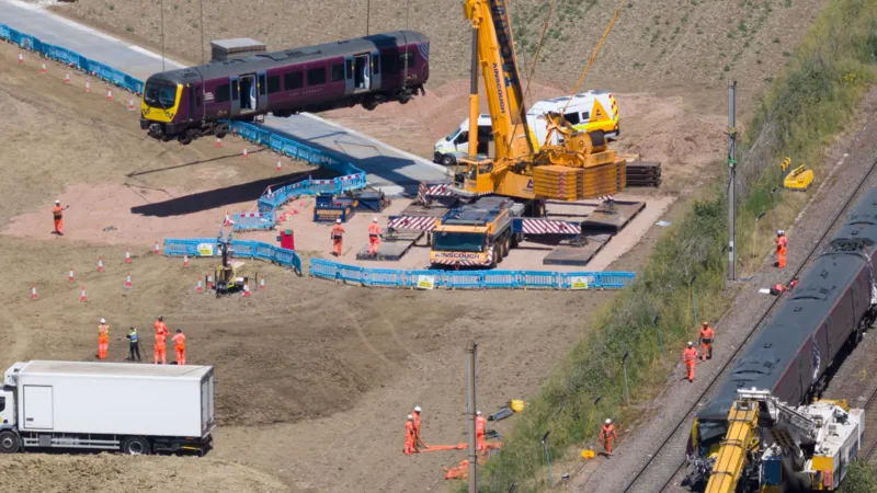 Train Manager Praised After Deadly East Midlands Railway Crash Near Bedford Emergency response at the crash scene A train manager has been praised for his quick thinking and bravery after a serious collision involving two East Midlands Railway services near Bedford. The crash happened on Friday at around 17:15 BST. One driver, Shaun Burton, died in the collision. Around 100 passengers were injured. Passengers said the train manager was hurt but still focused on safety. He used the radio to alert control rooms and helped coordinate an emergency response while checking on passengers. Heroic actions prevented further risk Representatives from the RMT Union said the train manager acted fast to prevent a second incident. He saw that one train had derailed and could block another line. He warned control staff immediately. He also placed circuit clips on the track system. This action forced signals to turn red and stopped other trains from entering the area. He then contacted the signaller to shut down movement on the line. Union officials said these actions may have stopped another train from hitting the damaged carriages. Witness accounts from passengers Passenger Mareks Grabovskis described the moment of impact. He said he had been in the last carriage after changing seats at the final moment. He said the crash threw him forward and he briefly lost consciousness. When he woke up, he saw injured passengers and panic inside the carriage. He also praised the train manager for staying calm and helping others even while injured himself. Tributes to driver Shaun Burton The driver who died, Shaun Burton, has been described as a respected member of staff. Local officials, including Blake Stephenson, shared condolences and highlighted the impact on his family and community. Sarah Conboy, leader of Huntingdonshire District Council, said Burton had served as a councillor between 2018 and 2022 and was well respected locally. Major rail disruption and repair work Recovery work is ongoing across the crash site. Officials say the damage is severe. A 600 metre section of track needs full replacement. Overhead electric lines also require removal and rebuilding. Heavy cranes are being used to lift damaged train carriages. Rail teams expect disruption between London and Bedford to continue for at least a week. Officials say access to the site is difficult, which is slowing recovery work. Medical response and passenger care A doctor who was travelling on one of the trains suffered fractures but still helped treat injured passengers at the scene. Emergency services later took the doctor to hospital. Passengers arriving at stations said staff have been providing support and updates as services slowly return.