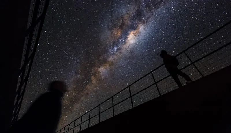 Atacama Desert’s Hidden Residencia: A Secret Base for Astronomers A building hidden in plain sight Deep in Chile’s Atacama Desert, a strange structure hides in the rocky landscape. From outside, the Residencia is almost invisible. Its entrance sits behind a plain door at the bottom of a ramp, blending into the dry terrain so well that most people walk past without noticing it. The surrounding land feels harsh and empty, with only rocky plains, distant mountains, and the occasional bird cutting through the dry sky. A sudden green world inside Stepping inside changes everything. The dry desert disappears, replaced by a cool, humid environment filled with life. Plants and trees grow in a central indoor garden, creating a green oasis in the middle of one of the driest places on Earth. At the heart of the space sits a bright blue swimming pool under a large dome. The air feels moist on the skin, a sharp contrast to the desert outside. A place that looks like a movie set The design of the Residencia gives it a cinematic feel. Its hidden corridors, open terraces, and unusual layout once attracted filmmakers. It even served as a filming location for a major spy movie finale, where dramatic scenes and explosions were shot across the building. Despite that dramatic past, the structure was not built for films. It was designed for science. Home for scientists and engineers Today, the Residencia serves as a private residence for astronomers and engineers. It cannot be booked by the public. More than one hundred rooms are reserved for staff working at nearby observatories. Most residents are involved with facilities like the Very Large Telescope on Cerro Paranal, only a short distance away. They live here while conducting research in one of the most extreme environments on the planet. Built for extreme conditions The European Southern Observatory operates the Residencia. The goal is simple: support scientific work in a remote desert where conditions are tough but the skies are perfect for astronomy. The design allows scientists to rest, recover, and continue their work without leaving the harsh environment that makes their research possible. A desert base like no other The Residencia sits between two worlds. Outside, there is silence, heat, and dry land. Inside, there is water, greenery, and calm. It feels more like a luxury retreat than a scientific base, yet its purpose is deeply practical. For the people working there, it becomes a temporary home while they study the universe from one of the clearest skies on Earth.
