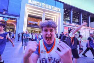 Knicks Bring Championship Fever Back to New York The New York Knicks have ignited a wave of excitement across New York City after reaching the NBA Finals for the first time since 1999. The team now leads the series 2 to 0 against the San Antonio Spurs, putting them just two wins away from a long awaited championship title. Game three takes place at Madison Square Garden, marking the first home game of the series. Fans across the city are treating it like a historic moment. High Security at Madison Square Garden Security has been significantly increased for the high profile game. Fans will face strict no bag rules along with airport like screening before entering the arena. US President Donald Trump is expected to attend the match, alongside New York Mayor Zohran Mamdani. The event has also attracted major celebrities including Timothée Chalamet, Ben Stiller, and Spike Lee. Authorities are preparing for heavy crowds and strong public attention due to the high stakes nature of the game. New York City Turns Orange and Blue The city has fully embraced the Knicks playoff run. Famous landmarks such as the Empire State Building and One World Trade Center have lit up in the team colors. Subway stations near Madison Square Garden now feature Knicks themed decorations. Local businesses across the five boroughs are offering special food deals, including orange and blue inspired snacks and desserts. Bars and public spaces are packed on game nights, with fans gathering to watch live broadcasts. Many streets near the arena have turned into unofficial celebration zones. Fans Celebrate a Historic Turnaround The Knicks success this season has surprised many supporters after years of poor performance. The team now stands one step away from a potential first championship since 1973. Even during away games in Texas, thousands of fans gathered in New York to celebrate wins. Some of these gatherings led to crowd control issues and several arrests as celebrations grew intense. Despite this, the atmosphere across the city remains highly energetic and focused on the possibility of a title win.