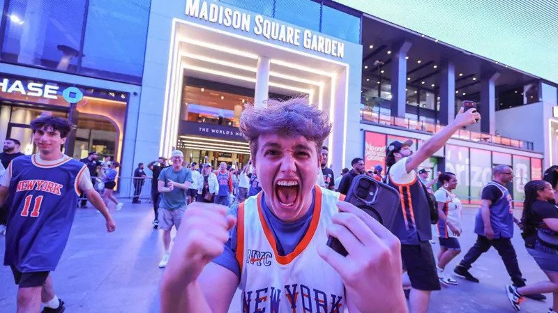 Knicks Bring Championship Fever Back to New York The New York Knicks have ignited a wave of excitement across New York City after reaching the NBA Finals for the first time since 1999. The team now leads the series 2 to 0 against the San Antonio Spurs, putting them just two wins away from a long awaited championship title. Game three takes place at Madison Square Garden, marking the first home game of the series. Fans across the city are treating it like a historic moment. High Security at Madison Square Garden Security has been significantly increased for the high profile game. Fans will face strict no bag rules along with airport like screening before entering the arena. US President Donald Trump is expected to attend the match, alongside New York Mayor Zohran Mamdani. The event has also attracted major celebrities including Timothée Chalamet, Ben Stiller, and Spike Lee. Authorities are preparing for heavy crowds and strong public attention due to the high stakes nature of the game. New York City Turns Orange and Blue The city has fully embraced the Knicks playoff run. Famous landmarks such as the Empire State Building and One World Trade Center have lit up in the team colors. Subway stations near Madison Square Garden now feature Knicks themed decorations. Local businesses across the five boroughs are offering special food deals, including orange and blue inspired snacks and desserts. Bars and public spaces are packed on game nights, with fans gathering to watch live broadcasts. Many streets near the arena have turned into unofficial celebration zones. Fans Celebrate a Historic Turnaround The Knicks success this season has surprised many supporters after years of poor performance. The team now stands one step away from a potential first championship since 1973. Even during away games in Texas, thousands of fans gathered in New York to celebrate wins. Some of these gatherings led to crowd control issues and several arrests as celebrations grew intense. Despite this, the atmosphere across the city remains highly energetic and focused on the possibility of a title win.