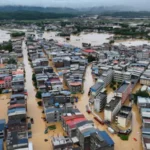 Typhoon Maysak Floods China After Heavy Rain Breaks Dam Wall Fast Rising Floodwaters Force Villagers to Escape Heavy rainfall from Typhoon Maysak caused severe flooding in Renhe village, located in Guangxi province, southern China. The rain started on Sunday, but residents did not expect the situation to become dangerous so quickly. Within hours, rising water entered homes and covered streets. By early Monday morning, floodwater reached knee level. Later, it completely covered the first floors of many houses. Residents said the water moved faster than expected. Many families escaped without taking food or important belongings because they had little time to react. A local resident named Zhou said the flood arrived suddenly and forced villagers to leave their homes immediately. Dam Damage and Rising River Levels Create Crisis Typhoon Maysak caused rivers across Guangxi to overflow. The powerful storm also damaged dam walls, increasing the danger for nearby communities. Thousands of people have left their homes as rescue teams continue operations in flooded areas. Several villages and parts of Nanning city faced serious water damage. Some residents waited on rooftops and called for help as floodwater surrounded their homes. Officials reported that the storm caused deaths and injuries across different regions. Rescue workers continue searching affected areas and providing emergency support. Extreme Weather Spreads Across China The storm also brought dangerous weather conditions to other parts of China. Hubei province experienced strong thunderstorms and rare tornado activity hundreds of kilometres away from Guangxi. The severe weather damaged communities and disrupted daily life for many residents. Chinese President Xi Jinping ordered emergency teams to increase rescue and relief operations. Authorities in Nanning warned that more heavy rain could make rescue work difficult. Climate Concerns Increase After Severe Storm Typhoons often bring heavy rainfall to China during this period of the year. However, experts say stronger extreme weather events have become a growing concern due to climate change. China’s agriculture sector, worth trillions of dollars, faces increasing risks from floods, storms, and changing weather patterns. Typhoon Maysak marks the first major storm to hit China during the 2026 typhoon season. Officials described the storm as sudden and powerful, with intense winds and short periods of extreme rainfall. More Storm Threats Ahead Weather experts are also monitoring another powerful storm, Super Typhoon Bavi, which could affect China’s eastern coastline later this week. Authorities continue to prepare emergency plans as communities recover from the destruction caused by Typhoon Maysak. The disaster highlights the growing need for stronger flood protection systems and faster emergency responses to protect people living in high risk areas.